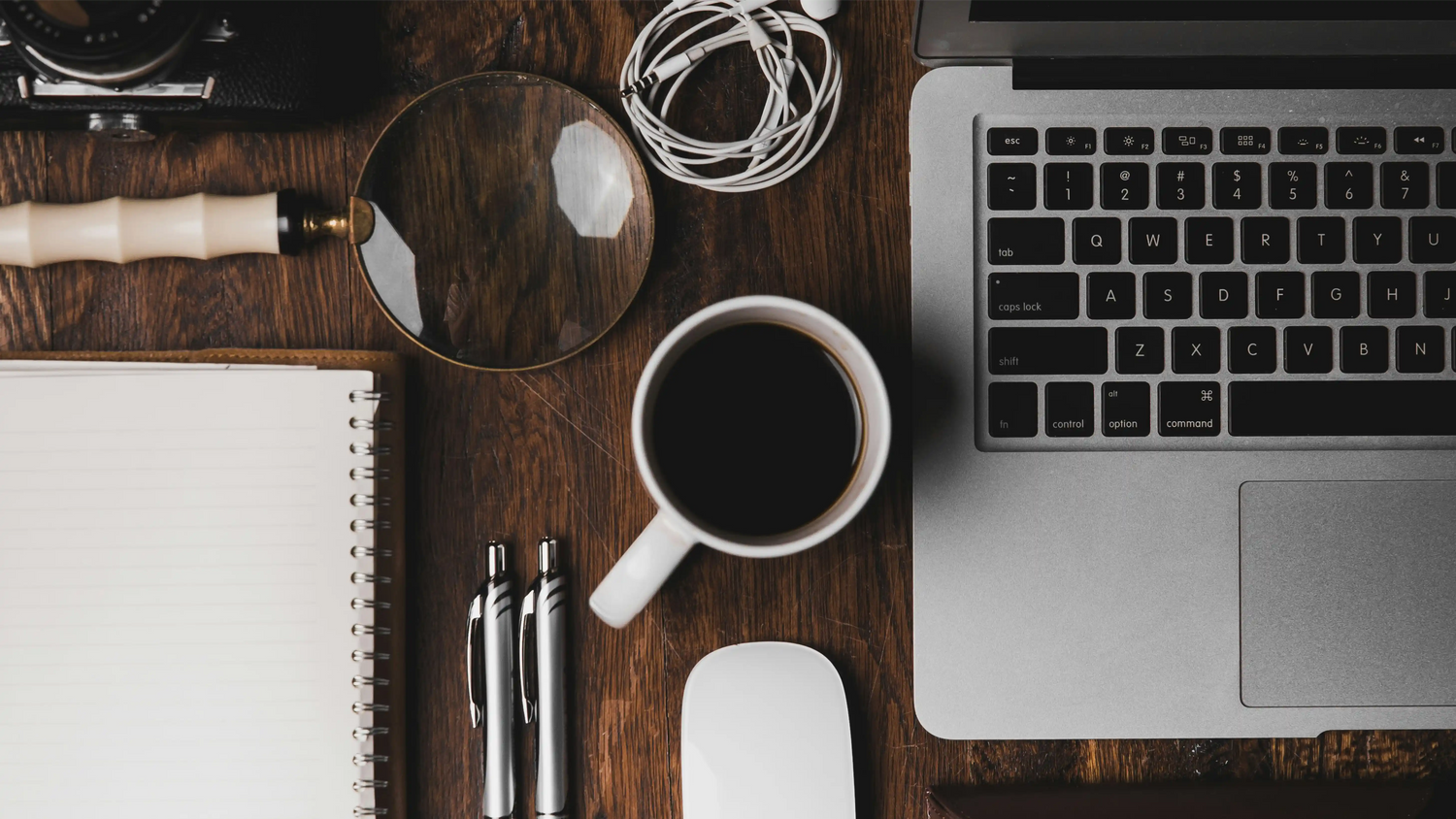 Minimalist office desk setup with laptop, coffee cup, white spiral notebook, magnifying glass, white earbuds and pens on wooden surface, captured from top view in moody lighting