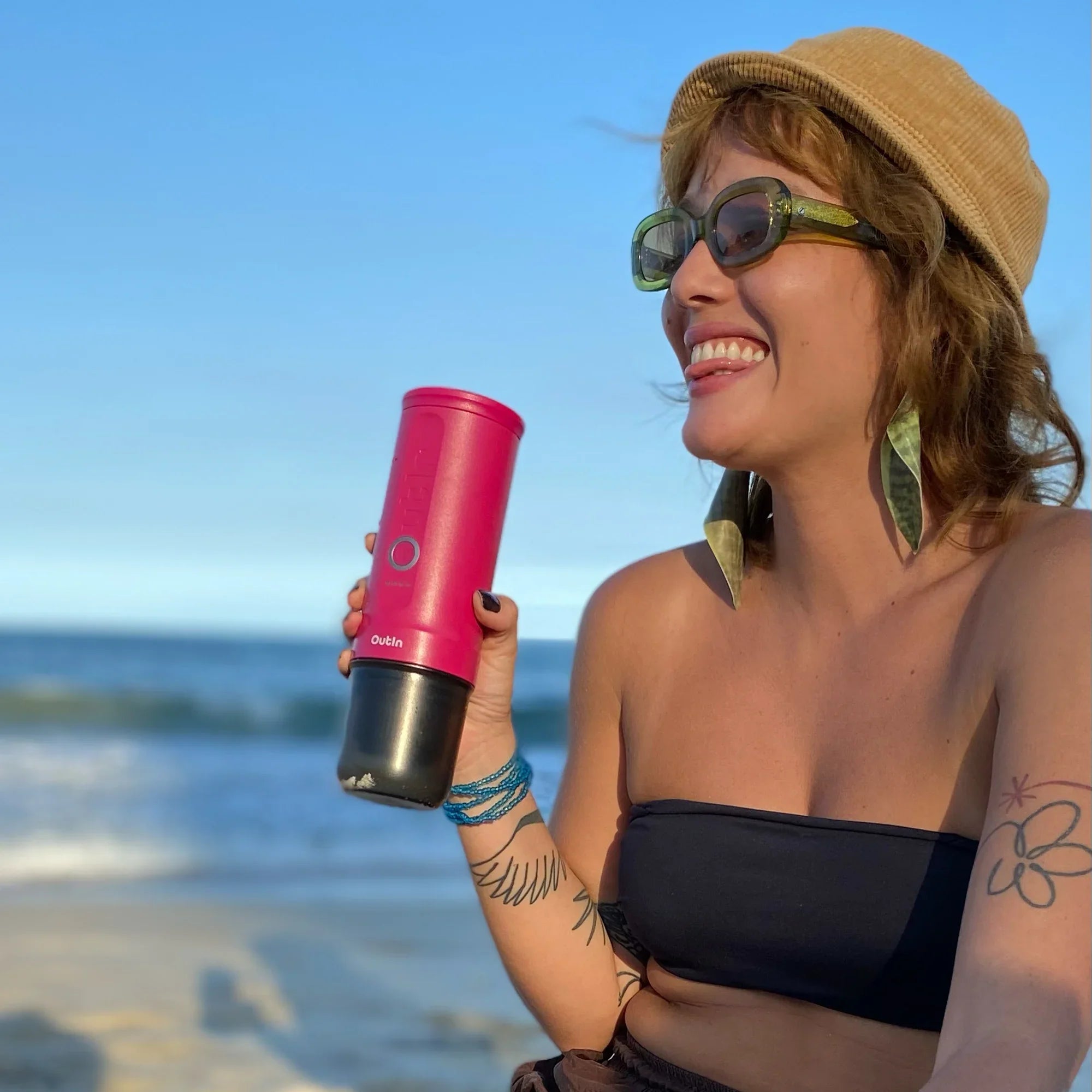 Smiling woman holding fuchsia Nano Portable Espresso Machine at the beach.